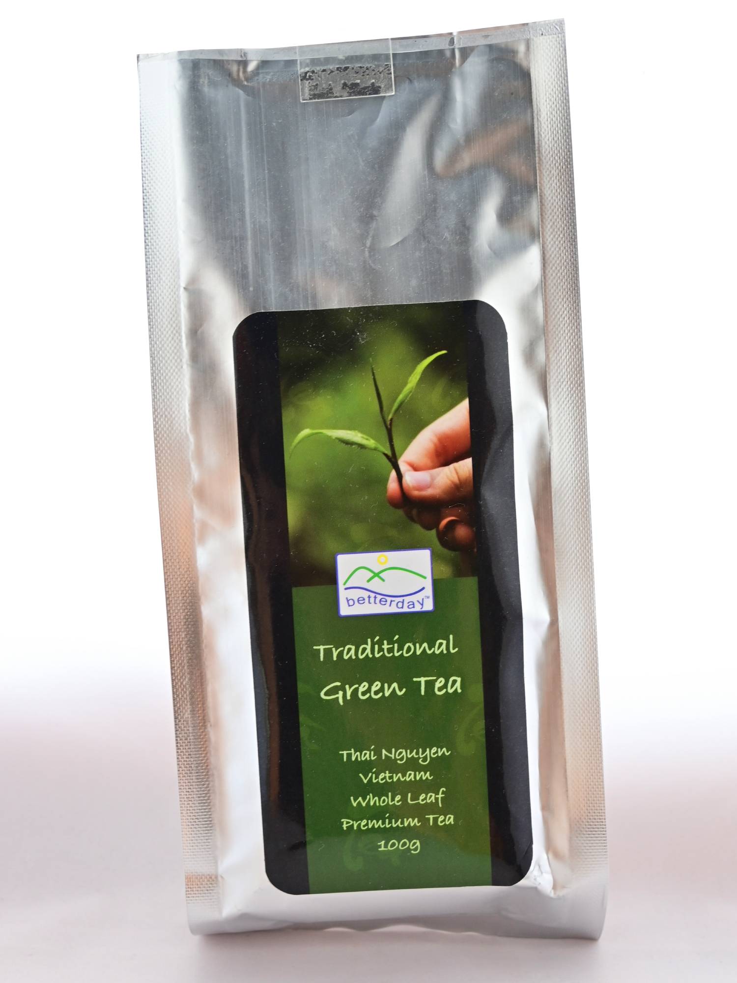 The plastic Che Xahn package is primarily a silver metallic color. In the center is a black label with green squares for text. A hand holds a tea bud. Beneath, text reads: "Traditional Green Tea. Thai Nguyen. Vietnam. Whole Leaf. Premium Tea. 100 grams."