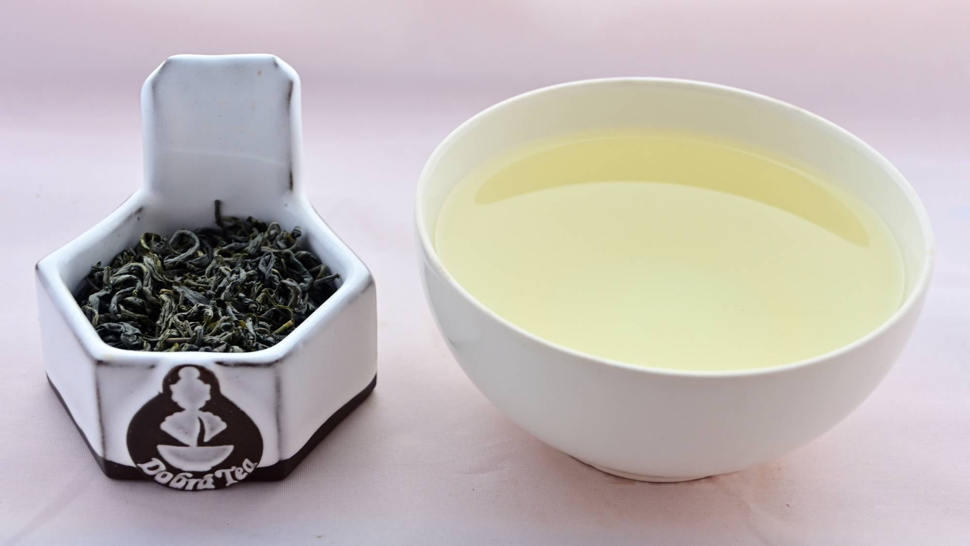 A side-by-side comparison of Che Xhan leaves and steeped tea. On the left, the leaves are a rich, dark green, and curl quite tightly while never fully rolling. On the right, the steeped liquid is a pale green.