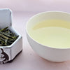 A side-by-side comparison of Lui'An Gaupian leaves and steeped tea. On the left, the leaves are long and narrow, and only barely curled into loose tubes. On the right, the steeped liquid is pale yellow.