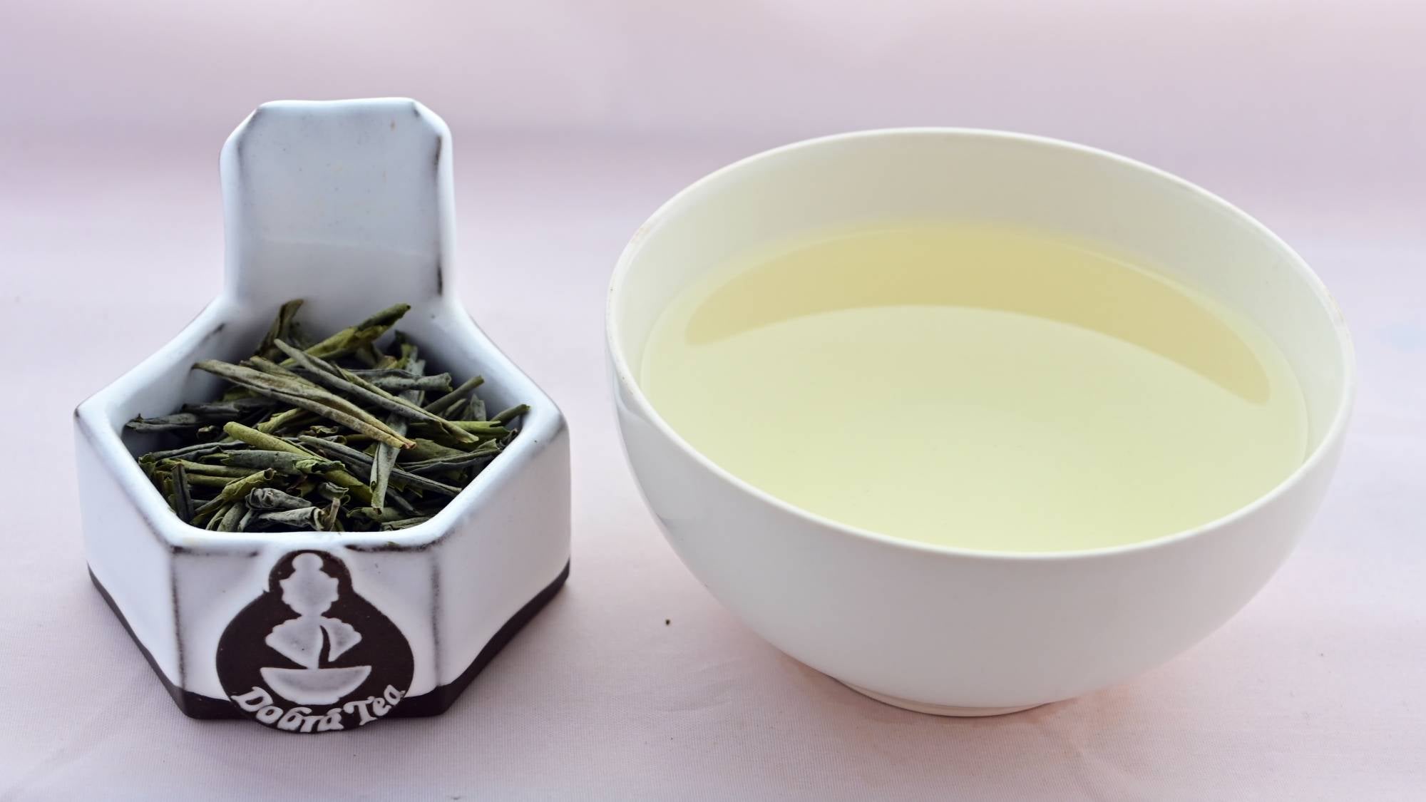 A side-by-side comparison of Lui'An Gaupian leaves and steeped tea. On the left, the leaves are long and narrow, and only barely curled into loose tubes. On the right, the steeped liquid is pale yellow.