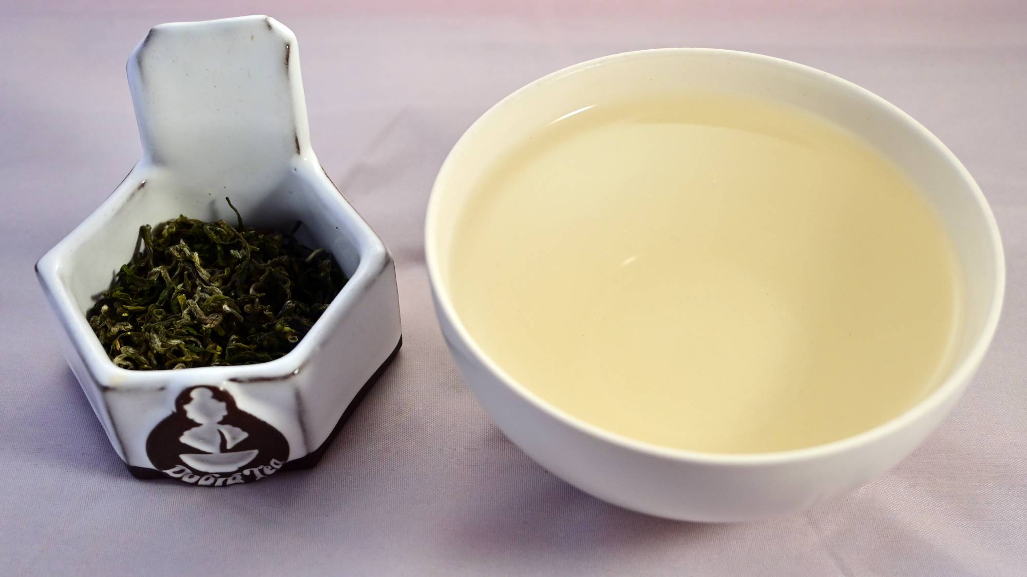 A side-by-side comparison of Putuo Fo Cha leaves and steeped tea. On the left, the leaves are a rich, dark green and are significantly curled with slight fuzz. On the right, the steeped tea is a pale green.