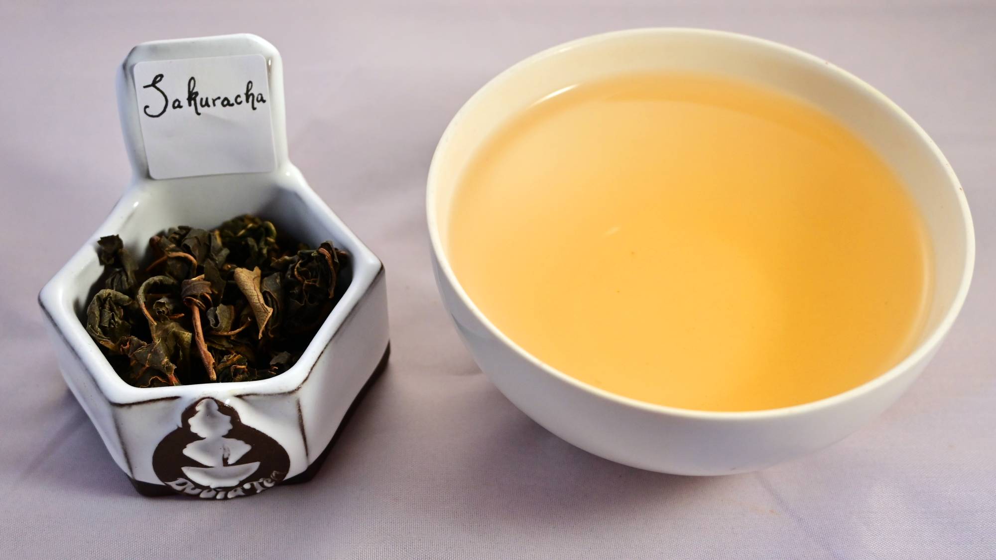 A side-by-side comparison of Sakuracha leaves and a cup of brewed tea. The leaves are whole and curled in on themselves. The steeped liquid is vibrant yellow.