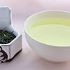 A side-by-side comparison of Shincha leaves and steeped tea. On the left, the leaves are predominantly dark green, long, flat, and narrow. On the right, the steeped liquid is a pale green.