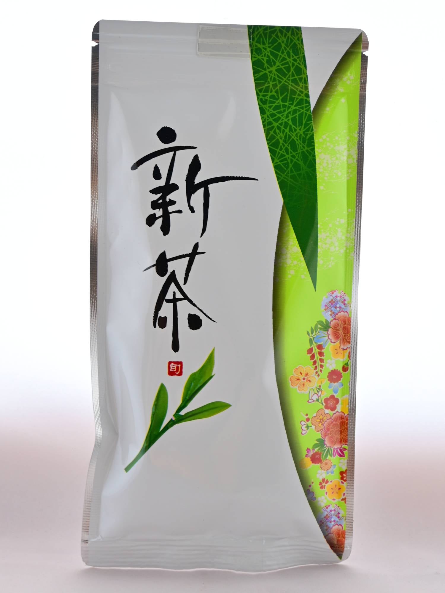 The Shincha package is mainly white, with green patterning on the right side. Inside one of the green semi-circles is a group of red and yellow flowers. On the left size of the plastic package are Japanese letters followed by a tea leaf.