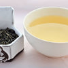 A side-by-side comparison of Zhu Cha leaves and steeped tea. On the left, the leaves are dark green, and resemble small, smooth pellets. On the right, the steeped liquid is a soft gold.