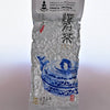 The plastic Ali Shan packaging is predominantly silver with a blue teapot in the bottom right. The package is rough and bumpy, characteristic of vacuum sealing around rolled tea leaves. A sticker at the top reads: "Dobra US LLC. 80 Church St. 05401 Burlington Vermont. Oolong Tea - Milky Oolong. Product of Taiwan. Net weight 1.7 oz."