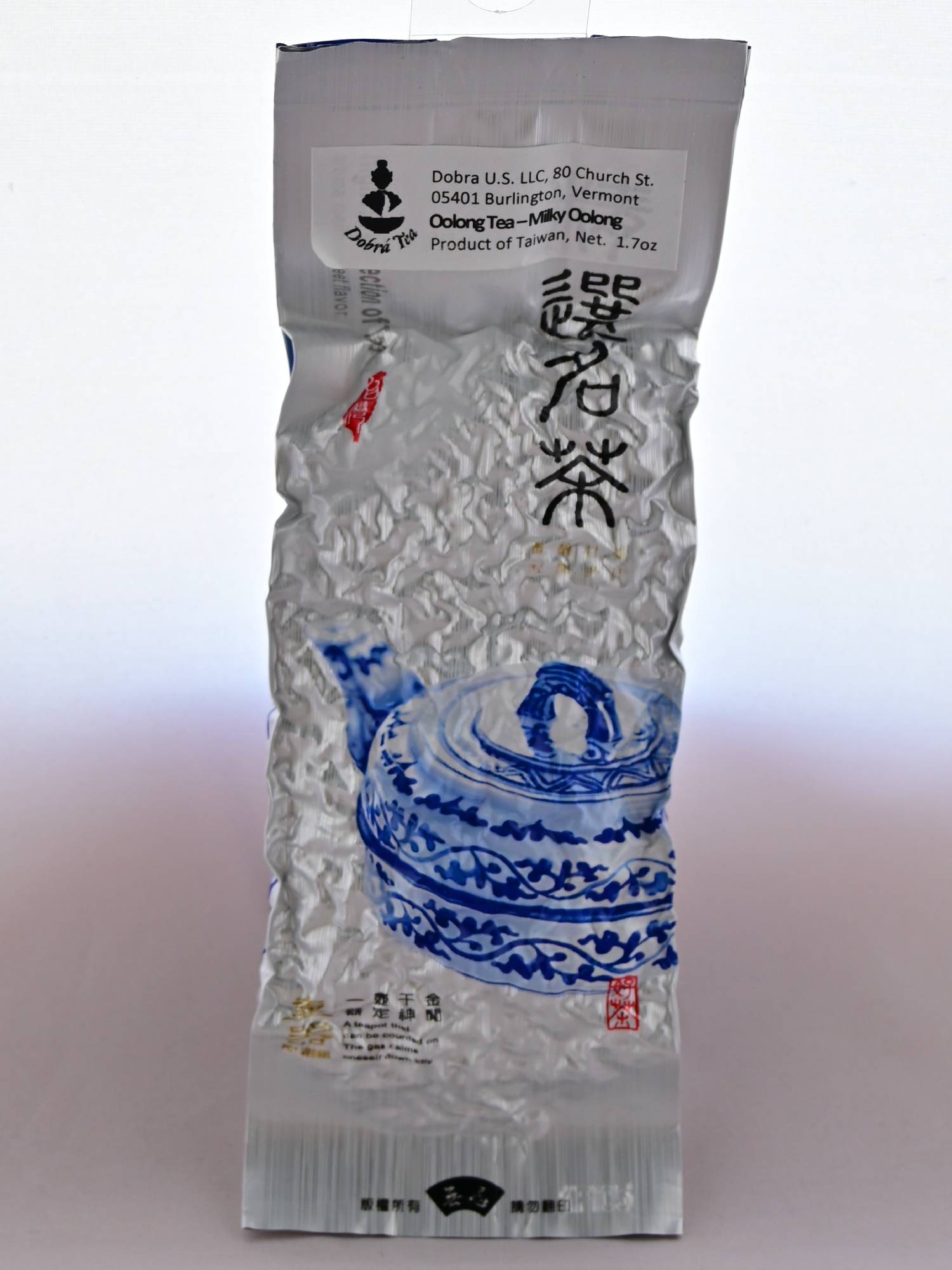 The plastic Ali Shan packaging is predominantly silver with a blue teapot in the bottom right. The package is rough and bumpy, characteristic of vacuum sealing around rolled tea leaves. A sticker at the top reads: "Dobra US LLC. 80 Church St. 05401 Burlington Vermont. Oolong Tea - Milky Oolong. Product of Taiwan. Net weight 1.7 oz."
