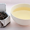 Crisp and curly tea leaves, rolled into long twisting shapes range from brown to pale green on the left. On the right, a cup of steeped Bai Hao tea is a pale peach color.