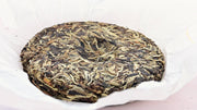 A full Bai Ya Bing Cha disk is featured against its white tissue-paper packaging. tea leaves ranging from light brown to a robust green are packed tightly into a disk shape with a small spherical divot in the middle.