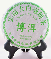 A Bai Ya Bing Cha tea disk sits on a black book holder. Chinese lettering in bright green runs around the package. The English translations are written underneath. "White Pu Ehr. Spring 2015. 200 grams."