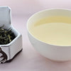 A side-by-side comparison of Bi Lou Chun leaves and steeped tea. On the left, the leaves range from bright green to dark green. They are twisted and curling, and appear slightly fuzzy. On the right, the steeped tea is a pale yellow color.