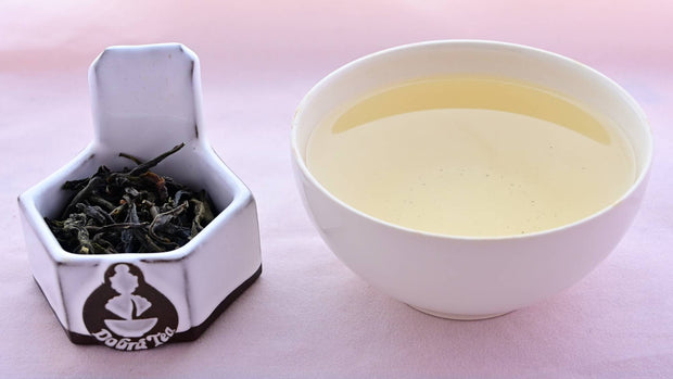 A side-by-side comparison of Burmese Wild Green leaves and steeped tea. On the left, the leaves are a rich dark brown with green undertone. On the right, the steeped tea is a soft yellow.