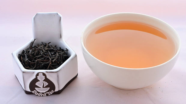 A side-by-side comparison of Ceylon Tiger River leaves and steeped tea. On the left, the leaves are a black color, and resemble small twigs. On the right, the steeped tea has a brown-red color.