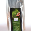 The plastic Che Xahn package is primarily a silver metallic color. In the center is a black label with green squares for text. A hand holds a tea bud. Beneath, text reads: "Traditional Green Tea. Thai Nguyen. Vietnam. Whole Leaf. Premium Tea. 100 grams."