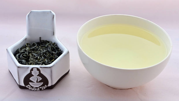 A side-by-side comparison of Che Xhan leaves and steeped tea. On the left, the leaves are a rich, dark green, and curl quite tightly while never fully rolling. On the right, the steeped liquid is a pale green. 
