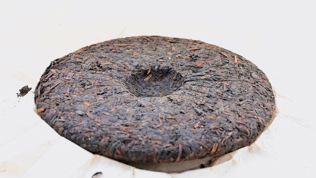 A full Chi Tse Bing Cha tea disk is shown against the white tissue paper wrapper. The leaves range in color from red-brown to dark brown, and are pressed into a circular disk with a spherical divot in the middle.