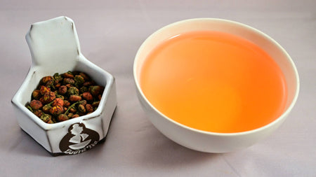 A side-by-side comparison of chrysanthemum buds and steeped tisane. On the left, the buds are tightly curled and orange with a bit of dark green dried leaf (resembling miniature pumpkins the size of a pen-point. On the right, the steeped tisane is dark orange.