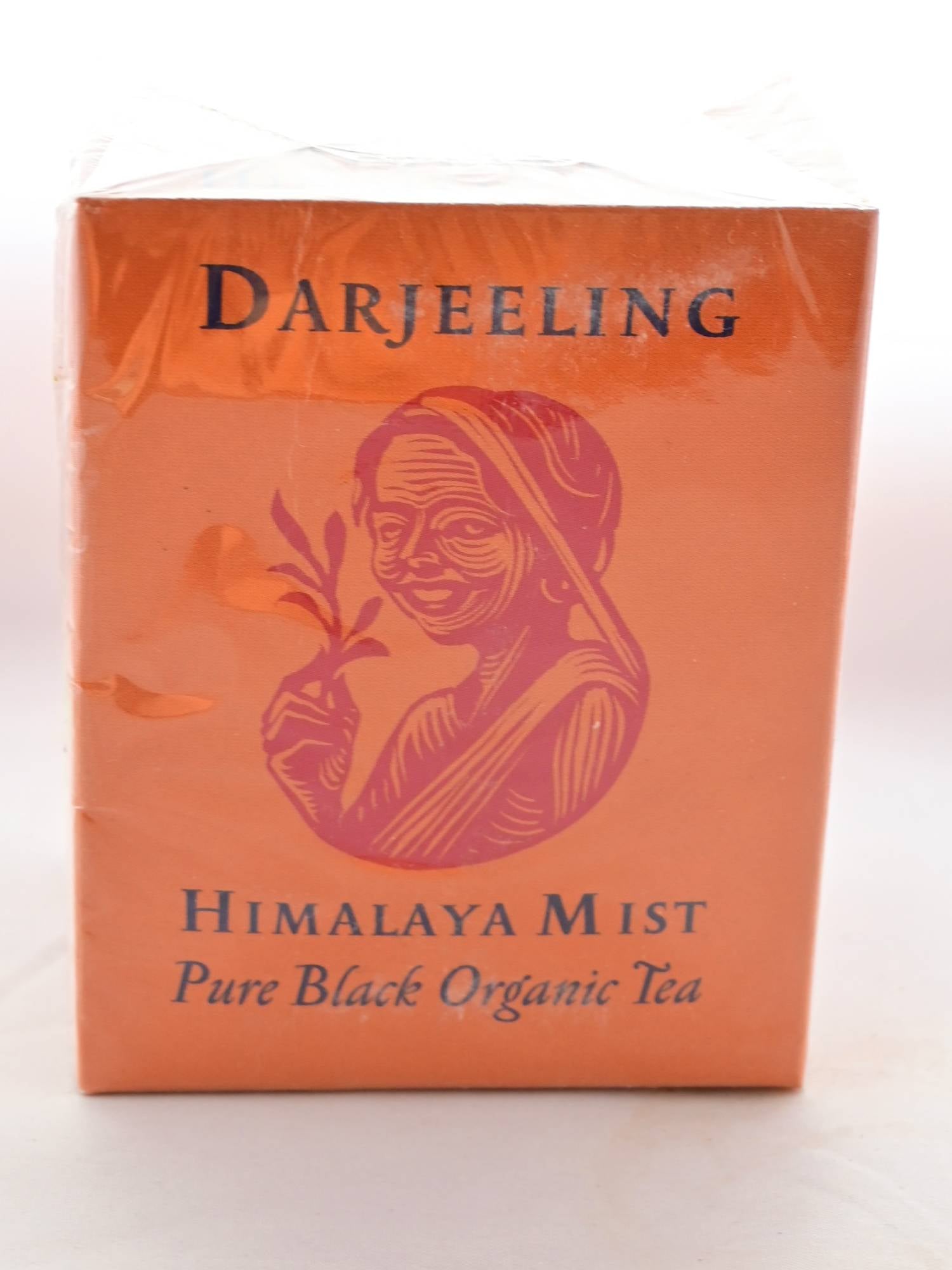 The Darjeeling Himalaya box is a squat orange cube. On the front, a line-drawing of a woman holding a tea plant smiles in red. Black text reads: "Darjeeling. Himalaya Mist. Pure Black Organic Tea."