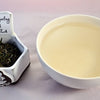 A side-by-side comparison of Darjeeling First Flush leaves and tea. On the left, the leaves are loosely curled and range in color from dark green to pale green. On the right, the steeped tea is a pale yellow.