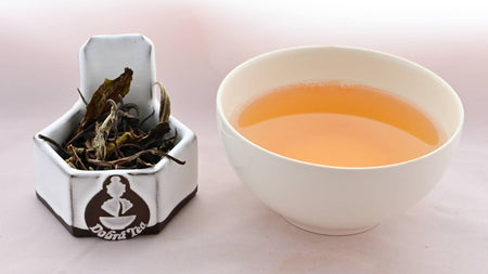 A side-by-side comparison of Dhara leaves and steeped tea. On the left, the leaves are tall, broad, and range in color from pale green to bright green. There are a few twig branches mixed among the leaves. On the right, the steeped liquid is a caramel color.