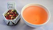 A side-by-side comparison of the Eastern Winds herbal blend and steeped tisane. On the left, the tea mixture prominently features hibiscus leaves and dried jasmine buds. On the right, the steeped tisane is dark orange.
