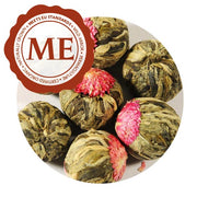 A close-up of several Jasmine Tea Flowers. A close-up of a single tea flour. A pink jasmine flower has been pressed into one side of the tea ball. Beneath the flower are a collection of pale green to dark green leaves, carefully packed and tied into a ball. They have a Meets EU Standards certification.