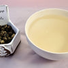 A bowl of rolled leaves next to a cup of prepared tea. The leaves are rolled into tight balls showing patches of light and dark color. The tea is pale yellow.