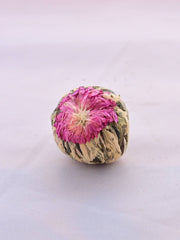 A close-up of a single tea flour. A pink jasmine flower has been pressed into one side of the tea ball. Beneath the flower are a collection of pale green to dark green leaves, carefully packed and tied into a ball.