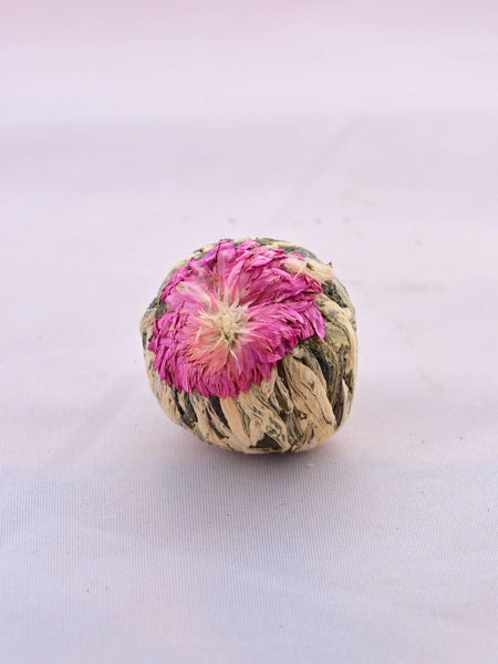 A close-up of a single tea flour. A pink jasmine flower has been pressed into one side of the tea ball. Beneath the flower are a collection of pale green to dark green leaves, carefully packed and tied into a ball.