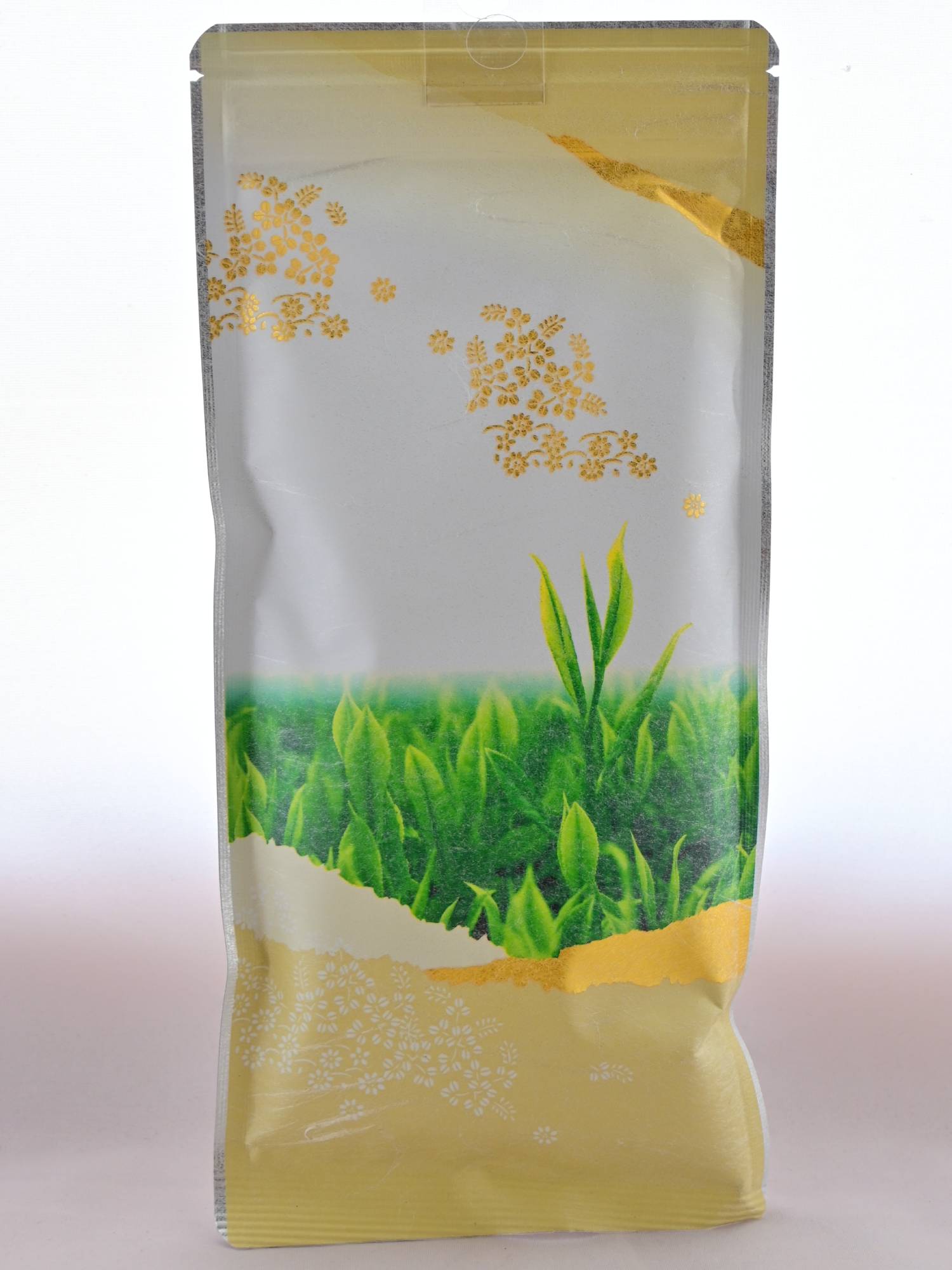 The plastic Kamacha packet has soft gold on the top and bottoms of it, with an image of the tops of tea bushes in the middle. There are golden flower fields and mountains painted along the edges. At the very top is a line of the resealable package closure.
