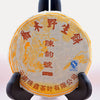 The Lao Shu Bing Cha disk is fully wrapped and displayed on a black book holder. The tissue paper cover is white, with yellow designs of a tree and someone looking at flowers on it. Red Chinese lettering circles the top, middle, and bottom. There is a blue stamp in the right-center.
