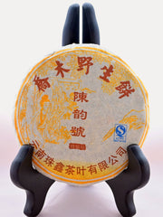 The Lao Shu Bing Cha disk is fully wrapped and displayed on a black book holder. The tissue paper cover is white, with yellow designs of a tree and someone looking at flowers on it. Red Chinese lettering circles the top, middle, and bottom. There is a blue stamp in the right-center.