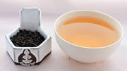 A side-by-side comparison of Lapsang Souchong leaves and steeped tea. On the left, the tea leaves are black and fit snugly together. On the right, the steeped tea liquid is pale orange.