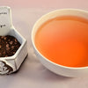 A side-by-side comparison of the Memories of Prague blend and steeped tea. On the left, the blend features tiny black tea pellets with crushed bits of dark chocolate. On the right, the steeped tea is a deep orange color.