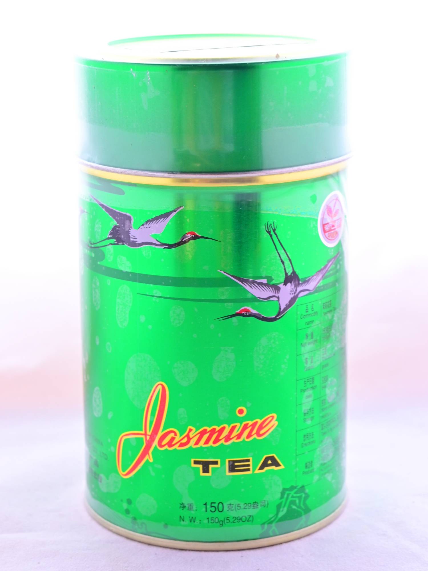 The metal Moli Hua Cha large tin is neon green, with storks flying along the top of the cylindrical canister. Toward the bottom, red and black text outlined in yellow reads: "Jasmine Tea. 150 g."