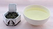 A side-by-side comparison of Nok Cha leaves and steeped tea. On the left, the leaves are medium-length and narrow, and appear to be a dark green color. On the right, the steeped liquid is a pale green.