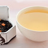 A side-by-side comparison of Plum Tea leaves and steeped tea. On the left, black tea leaves encircle nuggets of orange-colored dried plum. On the right, the liquid is a soft yellow color.
