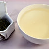 A side-by-side comparison of Putuo Fo Cha leaves and steeped tea. On the left, the leaves are a rich, dark green and are significantly curled with slight fuzz. On the right, the steeped tea is a pale green.