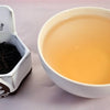 A side-by-side comparison of Qi Hong Mao Feng leaves and steeped tea. On the left, the leaves are small, tightly curled, and black. On the right, the steeped liquid is a soft orange.