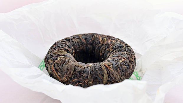 The Qing Tuo dome sits upside down in its white tissue paper wrapper. Leaves ranging from pale brown to dark brown are pressed together and curve inward.