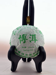 The Qing Tuo dome sits on a black book holder. It is wrapped in white tissue paper with green designs of flowers. Chinese letters wrap around the outside, with English translations on the inside reading: "Green Pu Ehr Tuo Cha. Spring 2015. 100 grams."