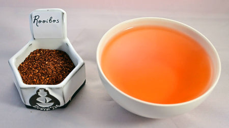 A side-by-side comparison of rooibos leaves and steeped tisane. On the left, the leaves are tiny, dust-like particles ranging in color from yellow to dark brown, with most being a bright red. On the right, the steeped tisane is a blood orange color.