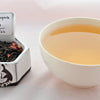 A side-by-side comparison of Casanova Rose Tea leaves and steeped tea. On the left, the tea leaves are a mix of tightly spiraled black tea with rose petals. On the right, the steeped tea is a pale orange color.