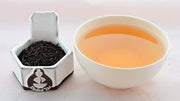 A side-by-side comparison of Rwandan Black leaves and steeped tea. On the left, teh tea leaves are small, tightly packed, and black. On the right, the liquid is a reddish brown mix.
