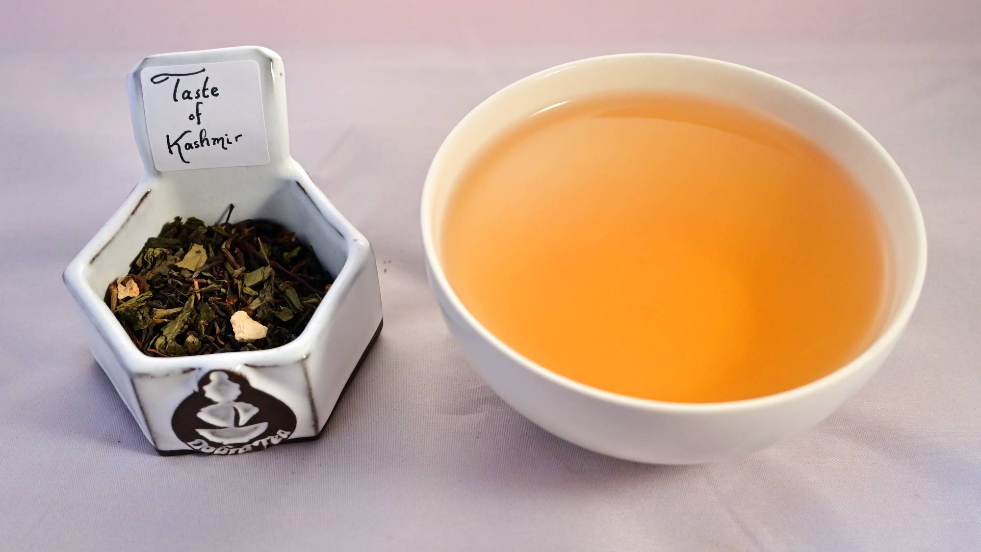 A side-by-side comparison of the Taste of Kashmir blend and steeped tea. On the left, the blend features a mixture of flat leaves, coiled leaves, and broken leaves which range in color from light brown to bright green, with bits of orange peel tossed in. On the right, the steeped tea is a soft orange.