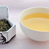 A side-by-side comparison of Touareg leaves and steeped tea. On the left, the leaves are a mixture of flat bright-green flakes and rounded, dark green pebbles. On the right, the steeped liquid is a soft yellow.