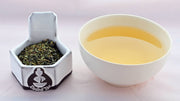 A side-by-side comparison of Touareg leaves and steeped tea. On the left, the leaves are a mixture of flat bright-green flakes and rounded, dark green pebbles. On the right, the steeped liquid is a soft yellow.