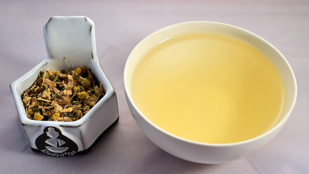 A side-by-side comparison of the Tummy Tamer blend and steeped tisane. On the left, the herbal blend prominently features chamomile and ginger root. On the right, the steeped tisane is a warm yellow color.