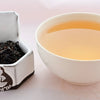 A side-by-side comparison of Vanilla Tea leaves and steeped tea. On the left, the tea leaves are black and slightly broken. On the right, the steeped liquid is a caramel color.