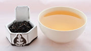A side-by-side comparison of Vanilla Tea leaves and steeped tea. On the left, the tea leaves are black and slightly broken. On the right, the steeped liquid is a caramel color.